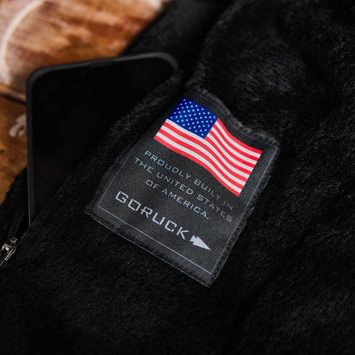 Close-up of a label on black fabric displaying the American flag and text Proudly Built in the United States of America above the GORUCK brand name. Next to the label, a smartphone is partially visible on a wooden surface, featuring GORUCK's Vest of Power - ToughDry® + Fleece commitment.