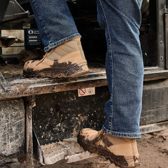 A person wearing the MACV-2 Safety Boot - Mid Top in tan by GORUCK and blue jeans steps onto a dirt-covered vehicle step. The background includes parts of the vehicle and a no-smoking sign.