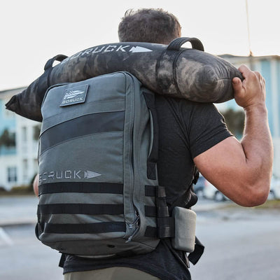 A person with a gray rucksack and a camouflage-patterned cylindrical bag slung over their shoulder strides confidently. Designed for heavy loads, both bags proudly display the GORUCK logo. The street, building, and clear sky provide an ideal setting for this urban adventure.