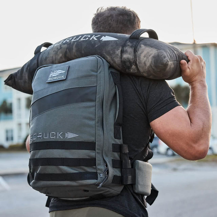 A person with a gray rucksack and a camouflage-patterned cylindrical bag slung over their shoulder strides confidently. Designed for heavy loads, both bags proudly display the GORUCK logo. The street, building, and clear sky provide an ideal setting for this urban adventure.