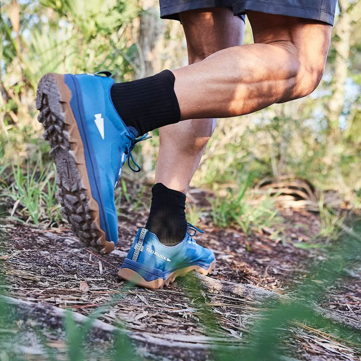 A person wearing GORUCK's Mackall shoes in Legion Blue + Gum and black socks walks through a forest path. The rugged soles, similar to those used in Special Forces training, provide excellent traction. The lush surroundings of grass and trees paint a vivid outdoor scene.