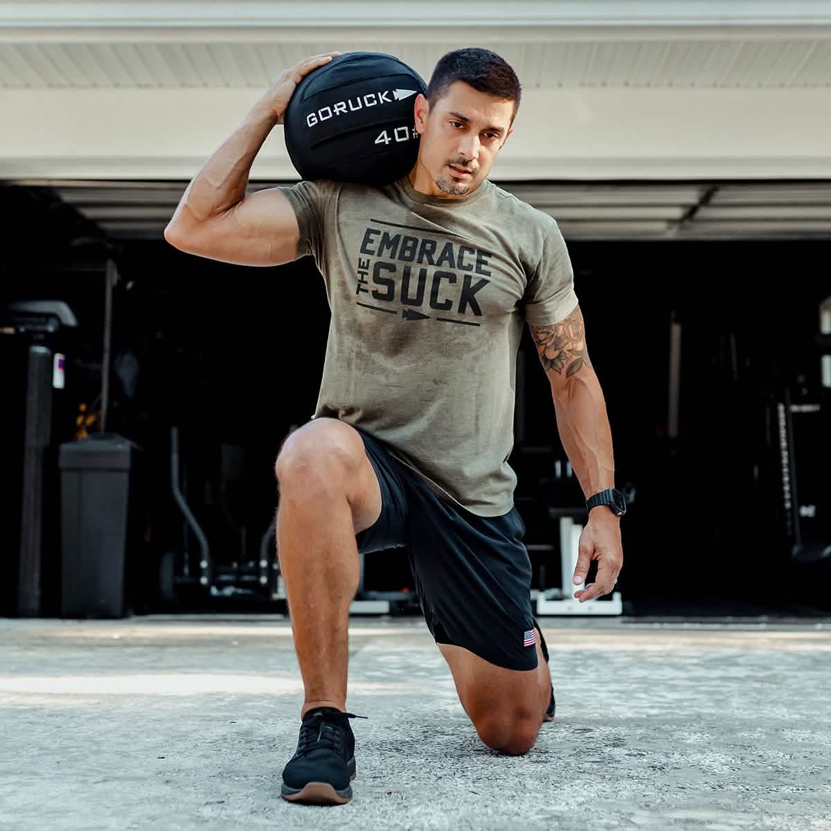 A man kneels on one leg in the driveway, with a GORUCK Sand Medicine Ball resting on his shoulder. He's dressed in a tan 