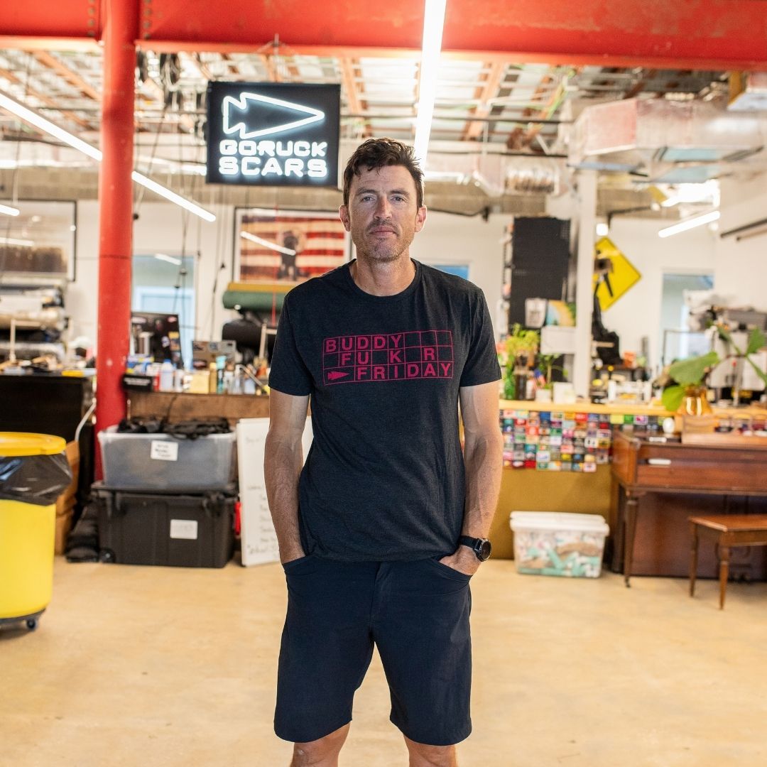 A man wearing a black t-shirt and shorts stands in an industrial workspace with a Buddy FCKR Friday sign behind him.