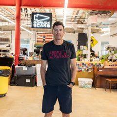 A man wearing a black t-shirt and shorts stands in an industrial workspace with a Buddy FCKR Friday sign behind him.