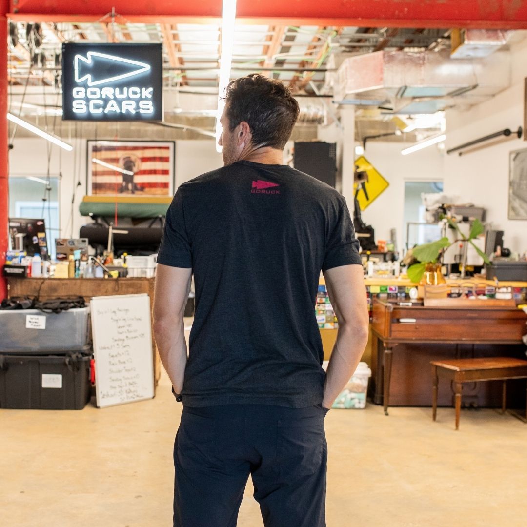 A man stands with his back to the camera in a workshop, beneath a GORUCK SCARS sign, wearing the Join or Die Tee - Tri-Blend—a shirt that channels the spirit of colonial unity inspired by Ben Franklin’s iconic cartoon.