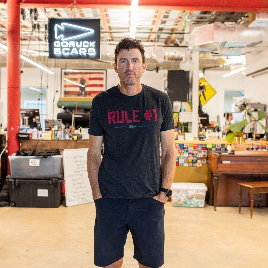 A man in a black “Rule 1” shirt stands in an industrial-style workspace with a GORUCK SCARS sign.