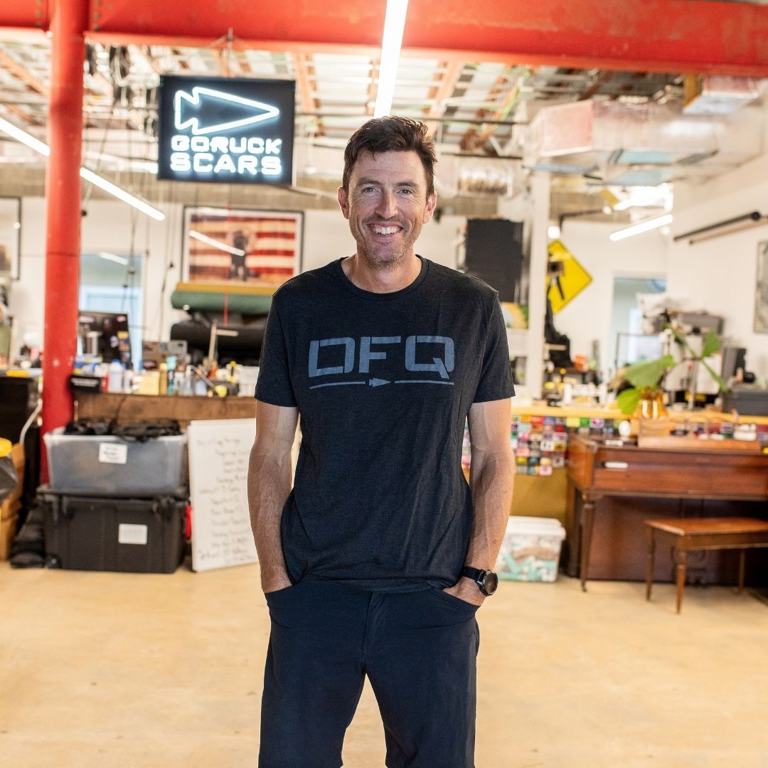 A man smiles while standing in a casual office with red beams, a piano, and a DFQ sign in the background.