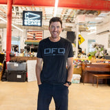 A man smiles while standing in a casual office with red beams, a piano, and a DFQ sign in the background.