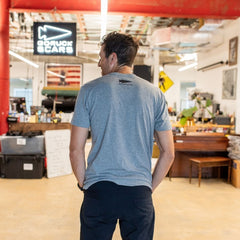A man wearing the Graphic T-Shirt - Built in the USA stands indoors, facing away in a workspace filled with equipment and a GORUCK sign.