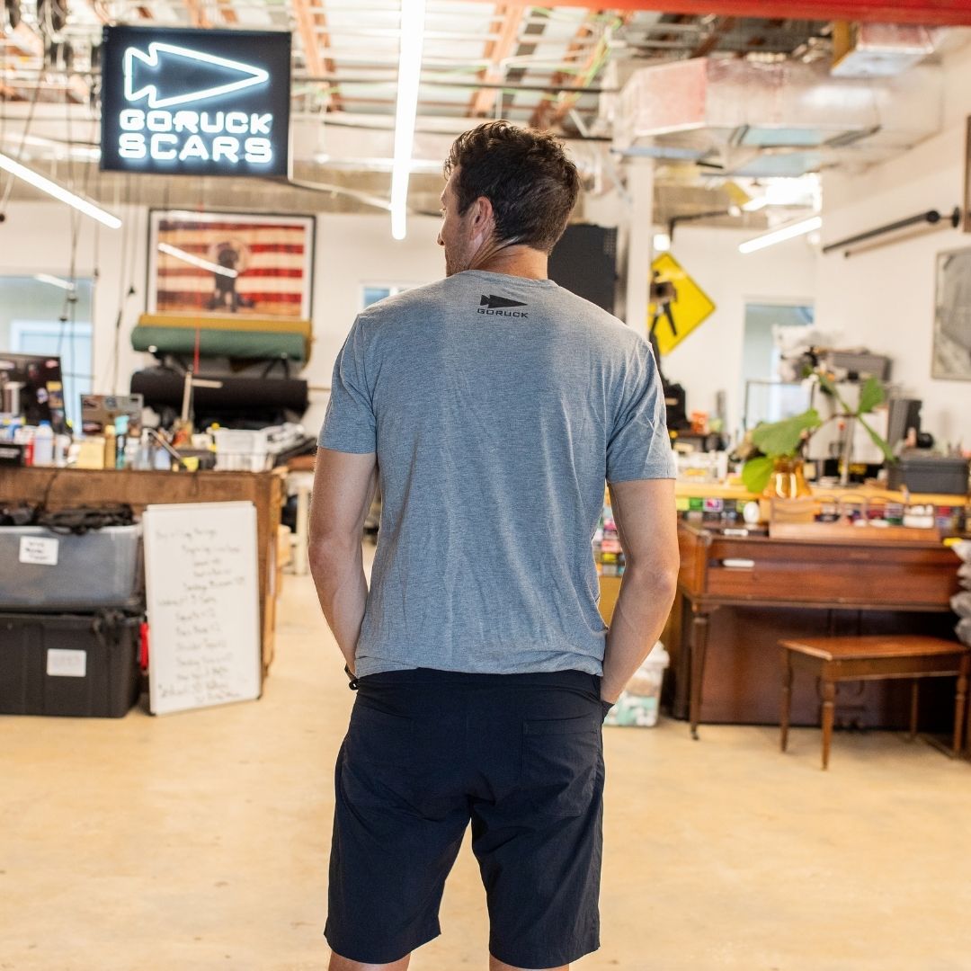A man in a gray t-shirt stands in a workshop, facing away, with an “In GOD We Trust” sign visible ahead.