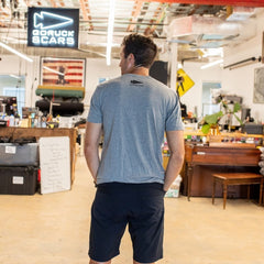 A man in a gray t-shirt stands in a workshop, facing away, with an “In GOD We Trust” sign visible ahead.