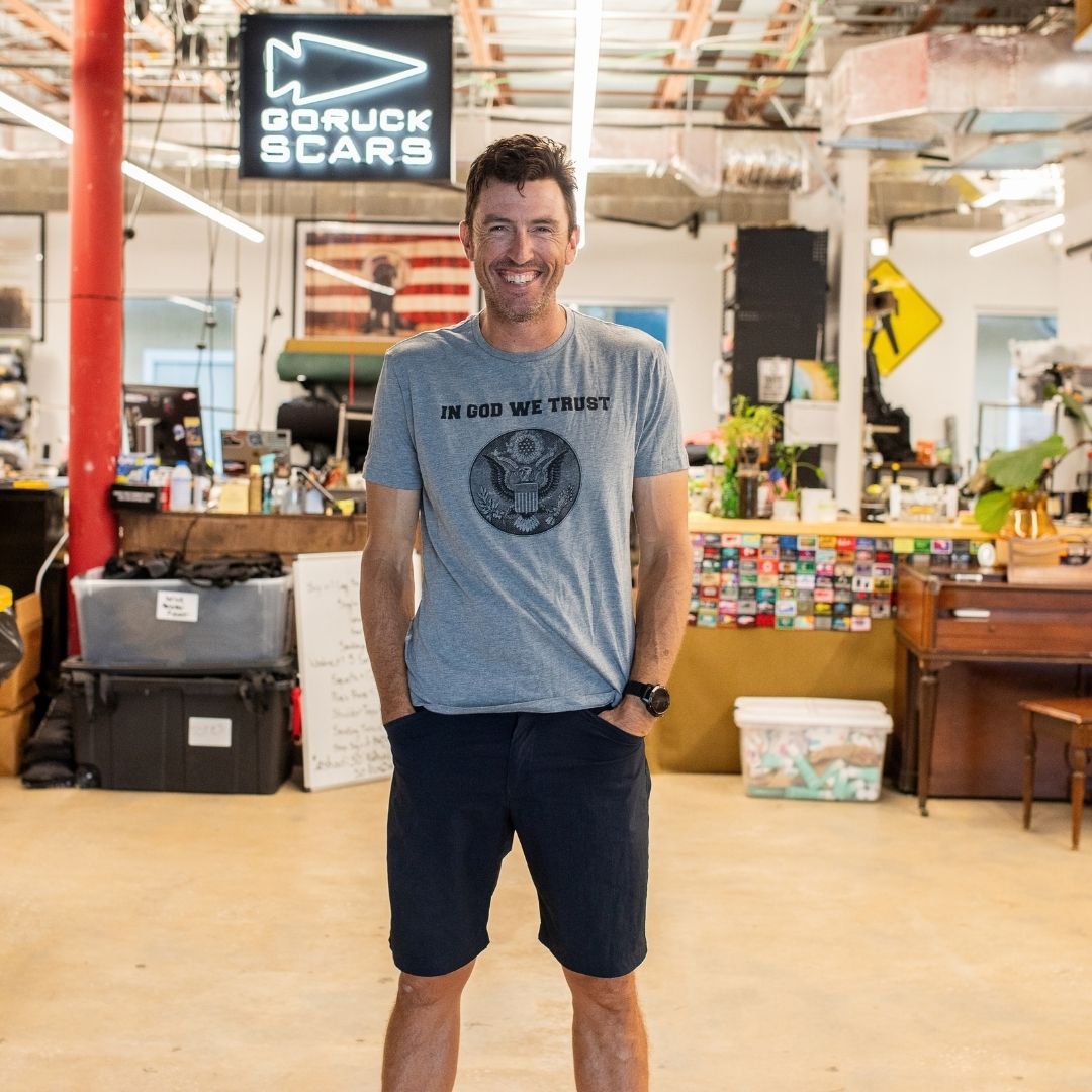 A man smiling in casual clothes stands in a workshop beneath a sign that reads “In GOD We Trust.”.