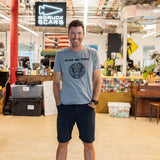 A man smiling in casual clothes stands in a workshop beneath a sign that reads “In GOD We Trust.”.