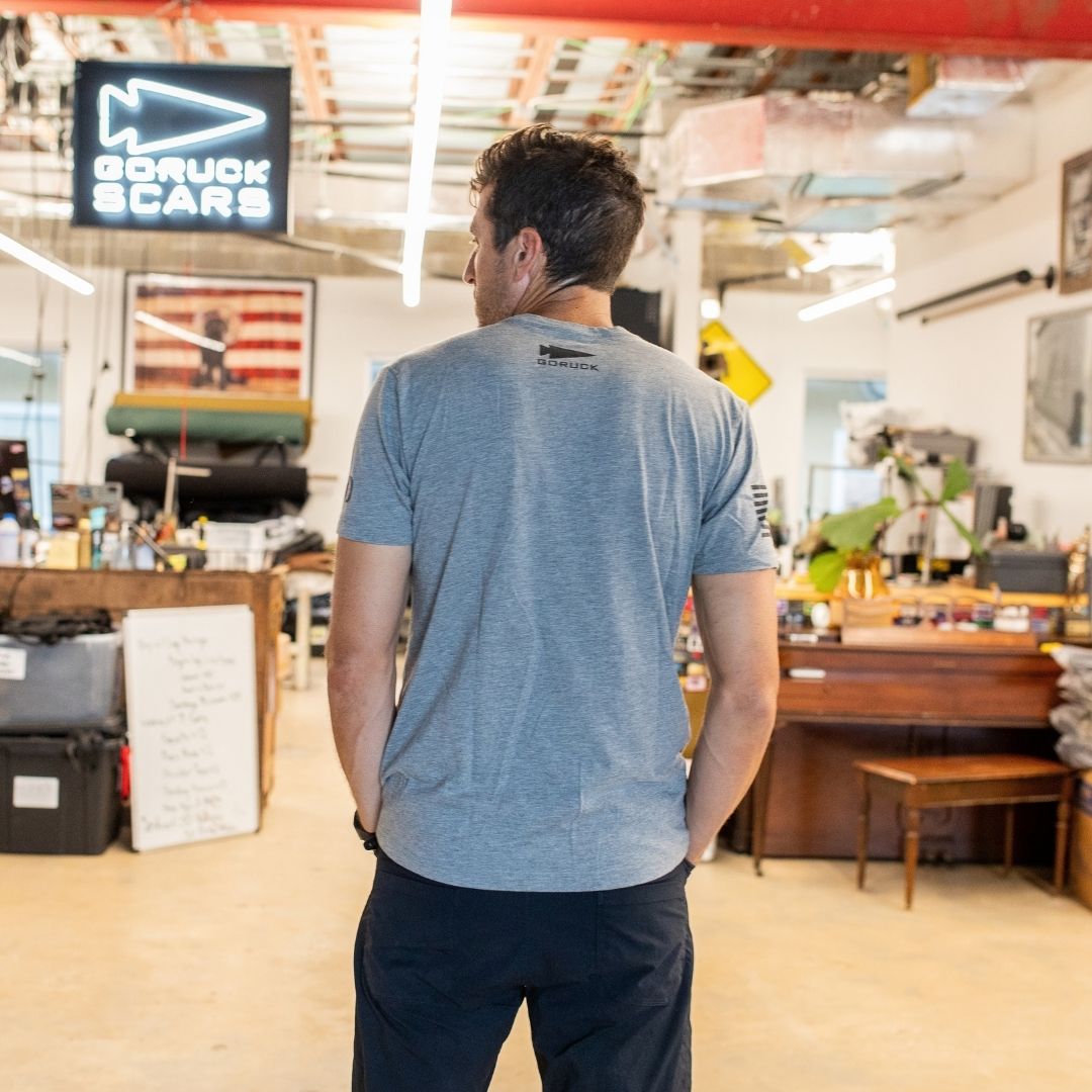 A man in a gray Join or Die Tee - Tri-Blend stands in a workshop, facing away, surrounded by equipment and a “GORUCK SCARS” sign—an atmosphere that echoes Ben Franklin’s era of craftsmanship and colonial unity, channeling the spirit of the legendary “Join or Die” cartoon.