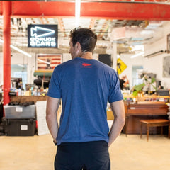 A man wearing the “America est. 1776” blue t-shirt stands indoors, facing away, with workshop equipment and signs in the background.