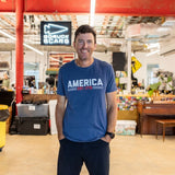 A man wearing the America est. 1776 shirt stands and smiles inside a casual office space.