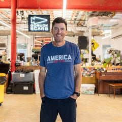 A man wearing the America est. 1776 shirt stands and smiles inside a casual office space.