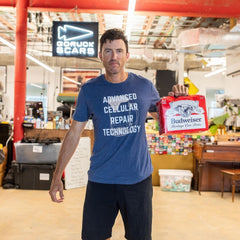 A man wearing a blue “ACRT” shirt holds a Budweiser box in a casual workspace.