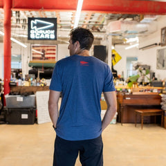 A man wearing the “Graphic T-Shirt - Built in the USA” stands with his back to the camera inside a workspace featuring red beams.