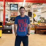 A man stands in a workshop wearing the Graphic T-Shirt - Built in the USA, featuring bold red text that reads “BUILT IN THE U.S.A.” on a blue background.