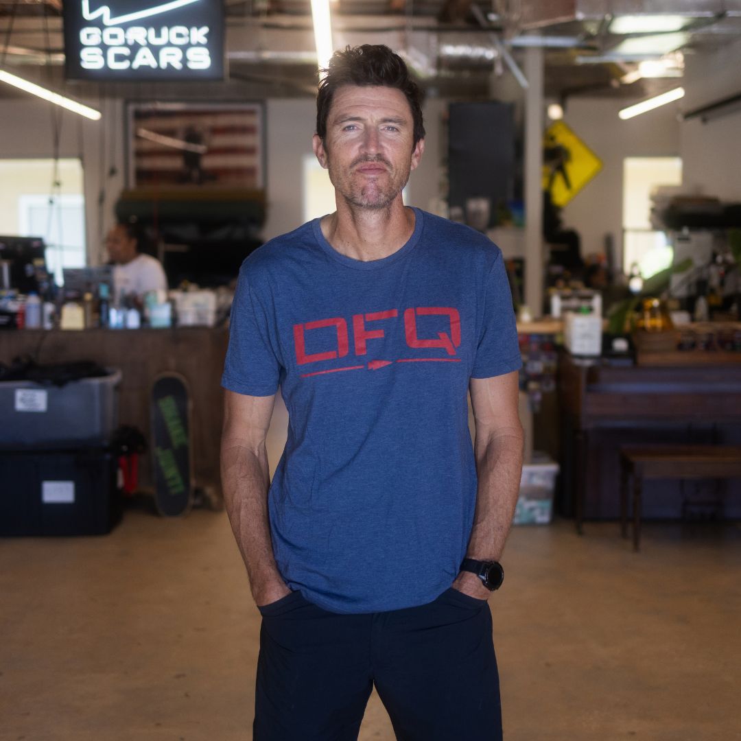 Wearing the DFQ TEE - TRI-BLENDX in blue, a man stands with his hands in his pockets inside a casual, industrial-style workspace. The bold “Don’t F*ing Quit” message on his shirt adds determination to the modern vibe.