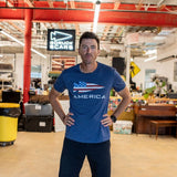 A man stands with hands on hips in a workshop, wearing the GORUCK Spearhead Tee - Stars & Stripes - Tri-BlendX, featuring the iconic spearhead emblem that channels a Special Forces spirit, with equipment in the background.