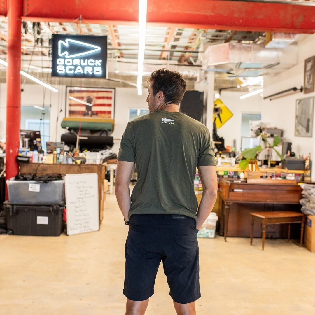 A man in a green “It Could Always Be Worse” premium tri-blend t-shirt stands with his back to the camera inside a cluttered workspace, showcasing quality apparel printed in the USA.