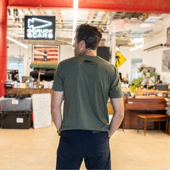A man in a green T-shirt stands indoors facing away, with a DFQ sign and an American flag in the background.