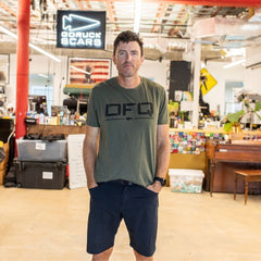 A man in a green T-shirt stands in an open office near a piano, facing the camera with his hands in his pockets, wearing DFQ.