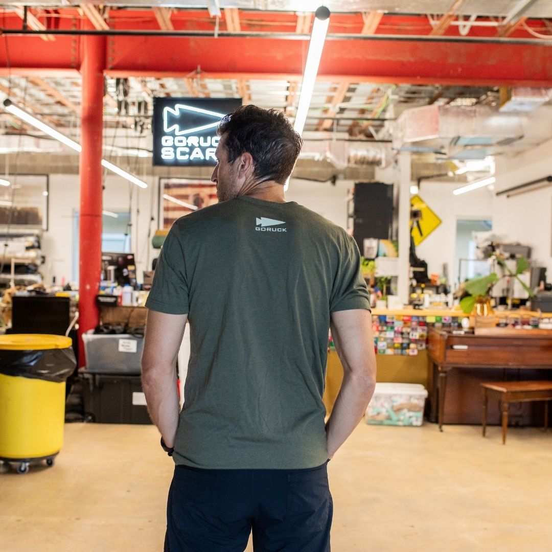 A man in a green shirt stands in an industrial-style workspace with bright lighting and red beams, featuring the “Easy Life Is Not For Us” product.