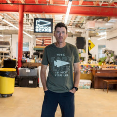 A man stands in a workshop, wearing the “Easy Life Is Not For Us” shirt with his hands in his pockets.