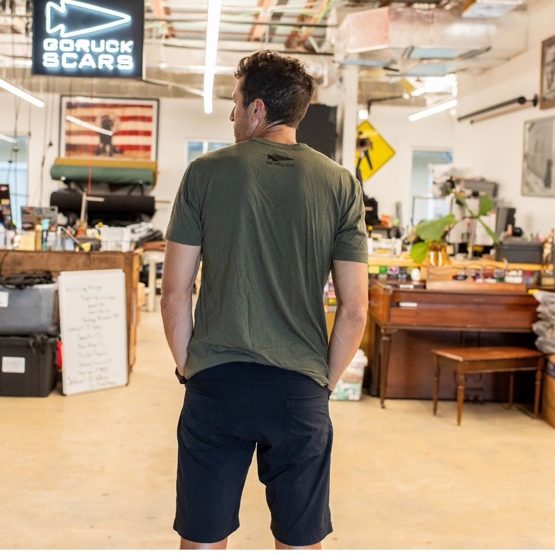 A man in an “In GOD We Trust” green shirt and black shorts stands in a spacious, cluttered workshop, facing away from the camera.
