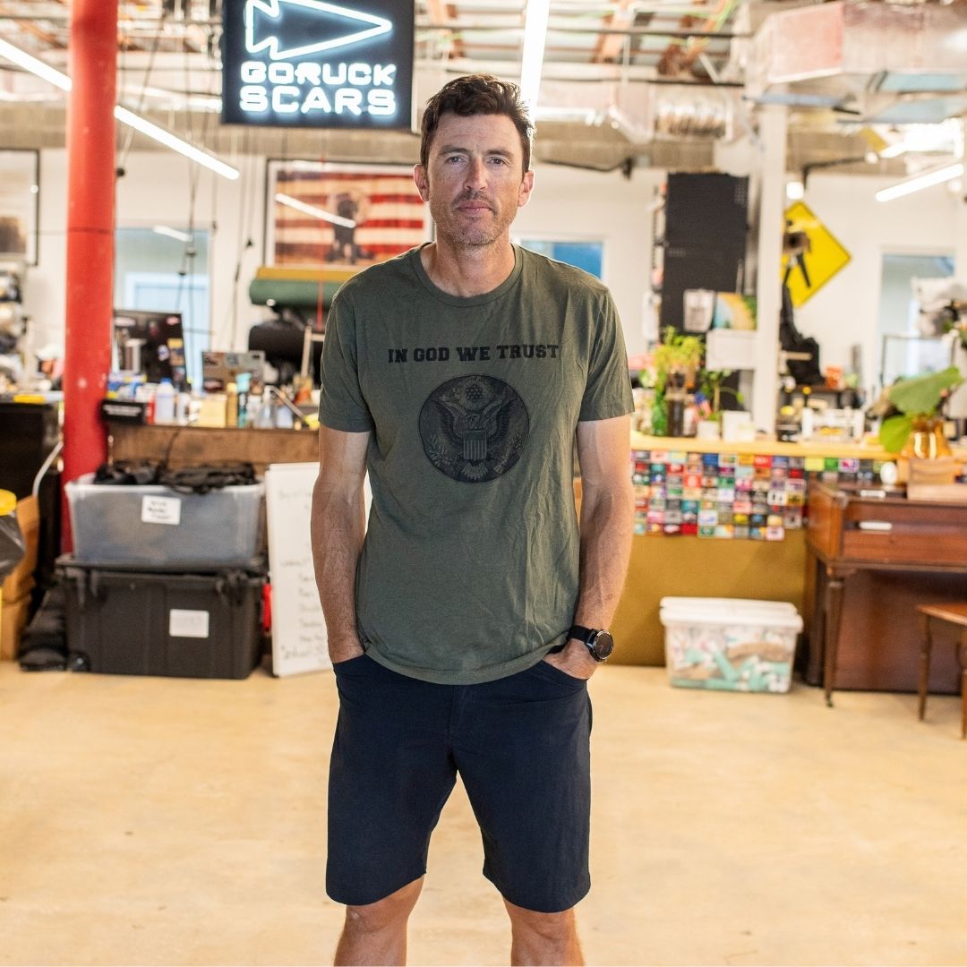 A man stands in an office wearing the “In GOD We Trust” t-shirt and shorts, looking at the camera.