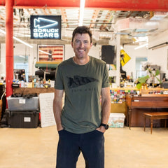 A man wearing the Join or Die Tee - Tri-Blend stands smiling in a workshop with red pipes and a “GORUCK SCARS” sign behind him, evoking the spirit of colonial unity first championed by Ben Franklin through his famous “Join or Die” cartoon.