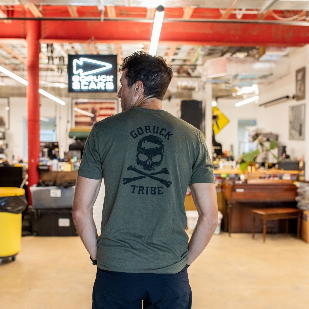 A man stands indoors wearing the GORUCK Tribe Tee - Tri-Blend in olive, featuring a skull and crossbones design on the back—a nod to the community spirit inspired by GORUCK Tribe and their legendary Challenge.