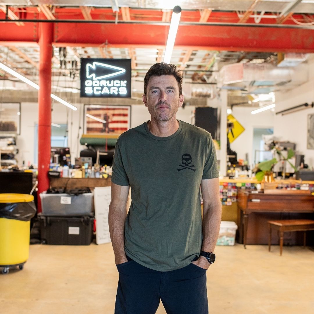 Wearing the GORUCK Tribe Tee - Tri-Blend, a man stands in a workshop with a “GORUCK SCARS” sign behind him, embodying the strong sense of community inspired by GORUCK Tribe.