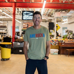 A man smiles in a workshop, wearing an “AmeRUCKa” shirt with his hands in his pockets; a busy workspace is visible behind him.