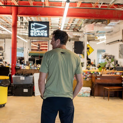 A man in a green T-shirt stands in a cluttered office beneath a sign reading “DFQ.”.