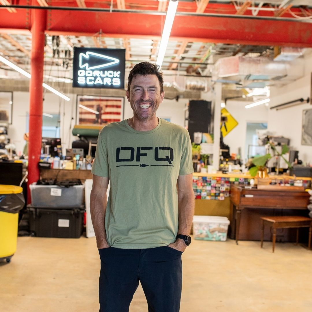 A man wearing a DFQ casual t-shirt smiles, standing in a bright, cluttered workspace with industrial decor.