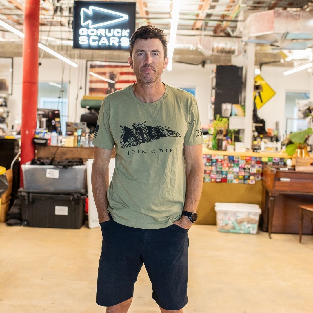 A man wearing the Join or Die Tee - Tri-Blend and navy shorts stands in an office with a “GORUCK SCARS” sign in the background, evoking the spirit of Ben Franklin’s iconic “Join or Die” cartoon and its call for colonial unity.