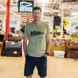 A man wearing the Join or Die Tee - Tri-Blend and navy shorts stands in an office with a “GORUCK SCARS” sign in the background, evoking the spirit of Ben Franklin’s iconic “Join or Die” cartoon and its call for colonial unity.