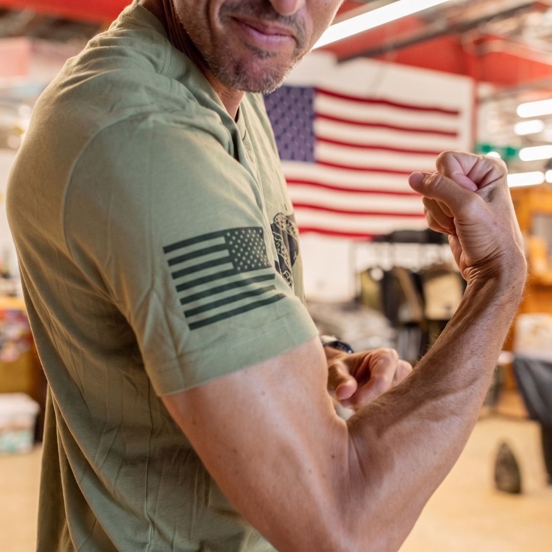 A man flexes his arm, wearing the Join or Die Tee – Tri-Blend in green, featuring a US flag design. The American flag is blurred in the background, capturing strength and patriotism inspired by Ben Franklin’s iconic message of colonial unity from the “Join or Die” cartoon.