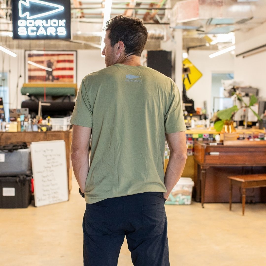 A man stands indoors facing away, wearing a green GORUCK Reflective Tee - Tri-Blend and dark pants in a casual, eclectic workspace.