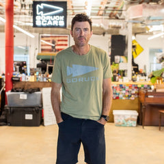 Man in a green GORUCK Reflective Tee - Tri-Blend stands in a casual, cluttered workspace with hands in pockets.