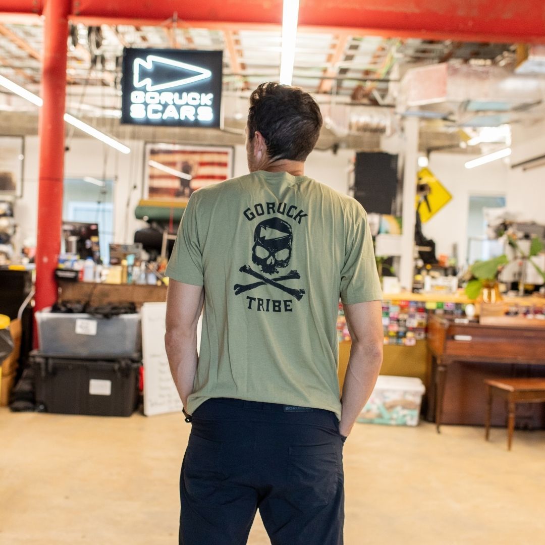 A man stands indoors, facing away, wearing the GORUCK Tribe Tee - Tri-Blend in green, featuring a skull and crossbones design—symbolizing the genuine community and spirit formed through every GORUCK Challenge.