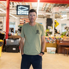 Wearing the GORUCK Tribe Tee - Tri-Blend, a man stands in a warehouse-style workspace accented by red pipes and a sign in the background, embodying the genuine sense of community that defines the GORUCK Tribe.