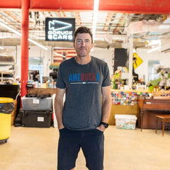 Man standing in warehouse, wearing “AmeRUCKa” t-shirt, with hands in pockets, smiling at camera.