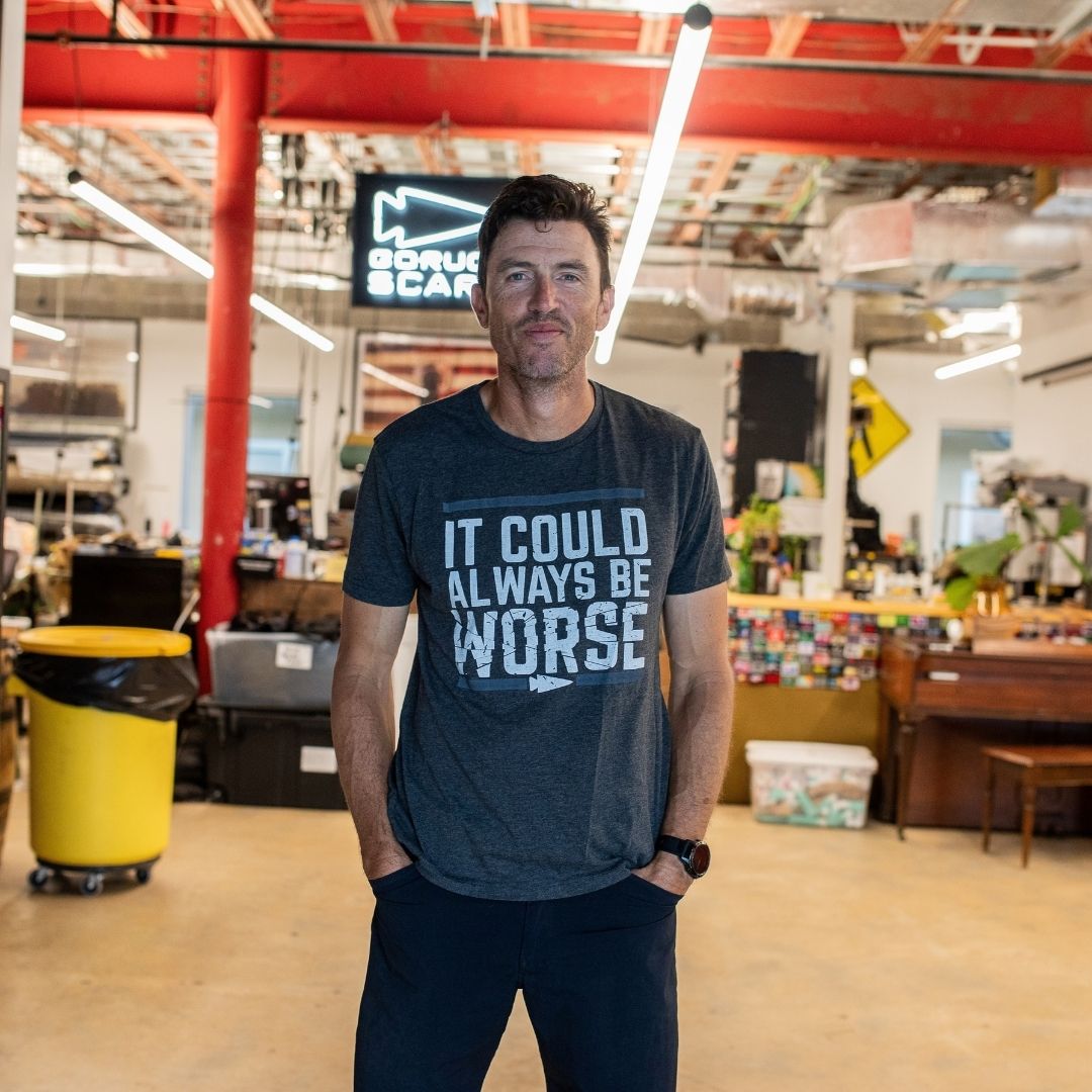 A man stands in a workshop with his hands in his pockets, wearing the “It Could Always Be Worse” t-shirt.
