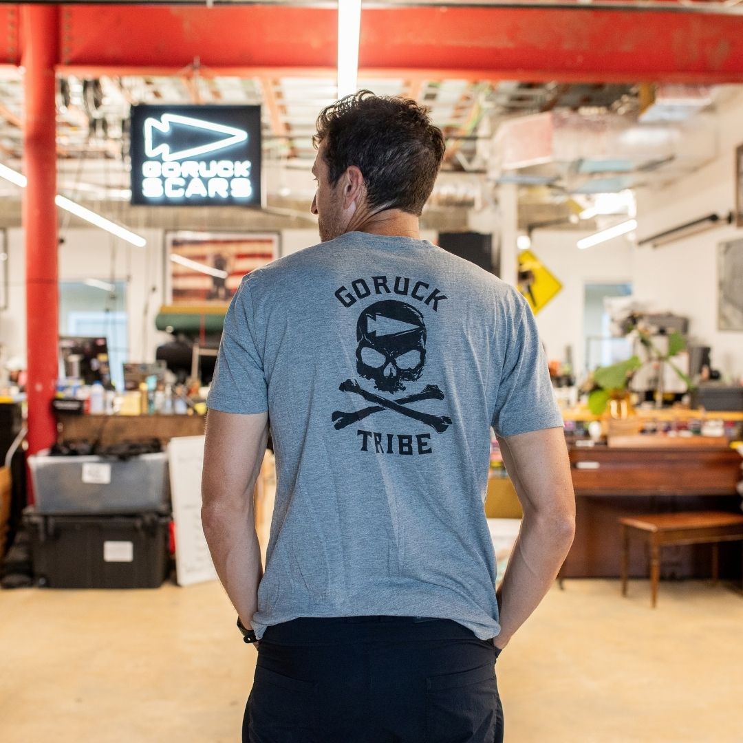 A man stands indoors wearing the GORUCK Tribe Tee - Tri-Blend in gray, featuring a skull and crossbones design on the back—ideal for those who value genuine community and embody the spirit of the GORUCK Challenge.