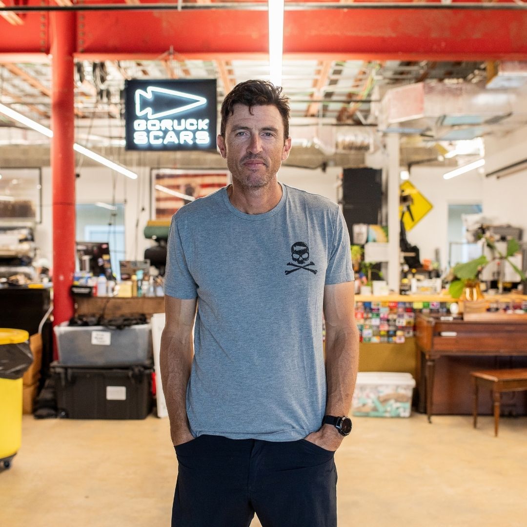 A man wearing the GORUCK Tribe Tee - Tri-Blend stands in a vibrant industrial workspace with a “GORUCK SCARS” sign, embodying the authentic community spirit of the GORUCK Tribe.